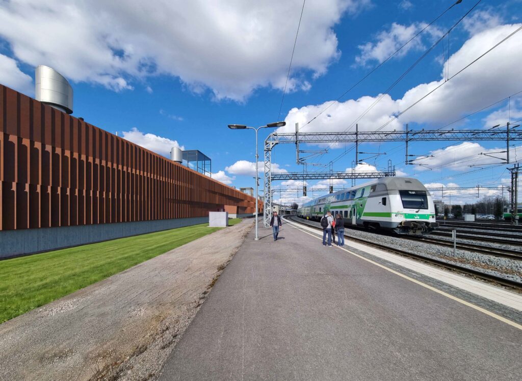 Valokuvaupote: Näkymä radan puolelta etelän suunnasta asemalaiturilta. HAMKin ruosteenvärinen kampus näkyy vasemmassa reunassa. On lähes pilvetön kesäpäivä. Ihmisiä kulkee asemalaiturilla, raiteilla kulkee juna.
