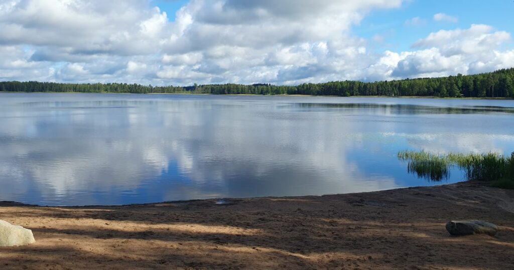 Lastujärven hiekkaisen uimarannan rannalta näkymä järven selälle. Puolipilvinen taivas heijastuu tyynen järven pinnasta.