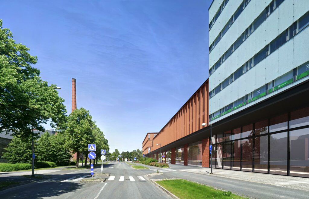 Valokuvaupote: Näkymä Eteläiseltä Asemakadulta pohjoiseen kohti uutta HAMKin kampusta. Rakennus on 3-4-kerroksinen, julkisivu on ruosteen väristä metallia ja ikkunapintaa. Etualalla näkyy olemassa oleva toimistotorni, jonka julkisivussa on vaaleaa lasitusta. On lähes pilvetön kesäpäivä.