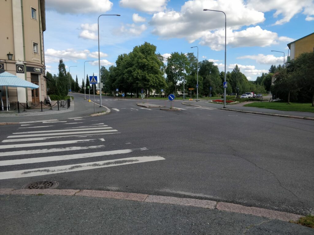 Riihimäen Kauppakadun ja Valtakadun risteys kuvattuna vihreän puistoalueen suuntaan. Risteyksen molemmilla puolilla näkyy rakennuksia.