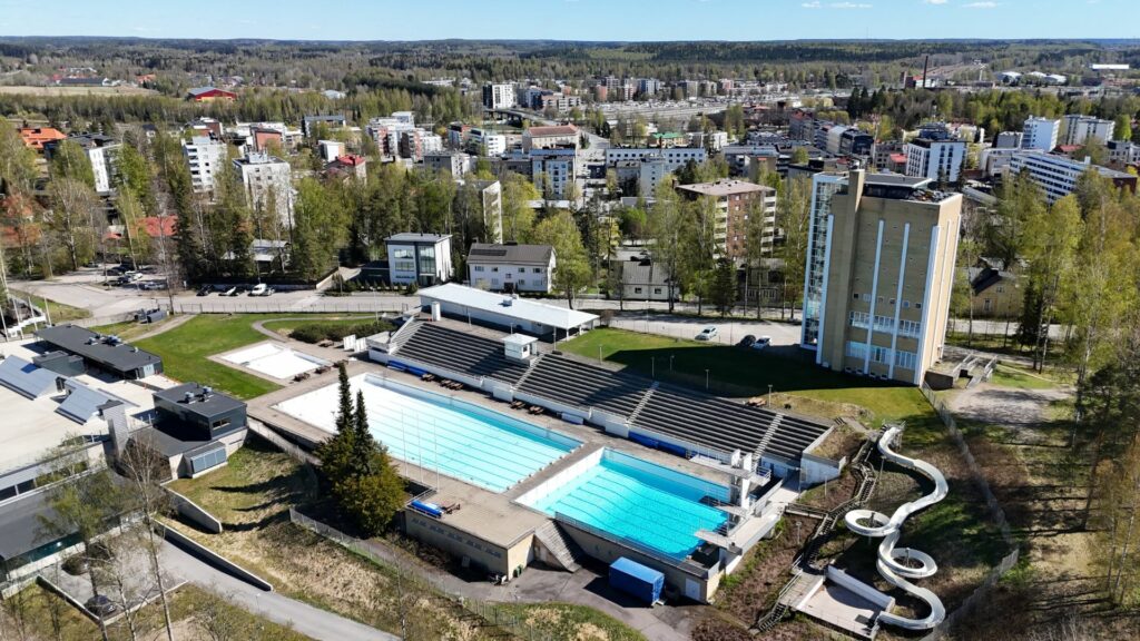 Maauimalan kaksi isoa allasta ilmasta kuvattuna. Vieressä korkea tornitalo sekä ympärillä kaupunkimaista tiheästi rakennettua maisemaa.