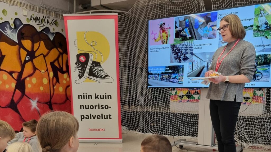 Nainen ison näytön edessä puhumassa. Etualalla lapsia. Rollup, jossa lenkkarit sekä teksti Niin kuin nuorisopalvelut.