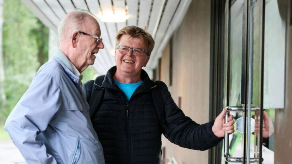 Kaksi vanhempaa miestä, joista toinen avaamassa toiselle ulko-ovea hymyillen.