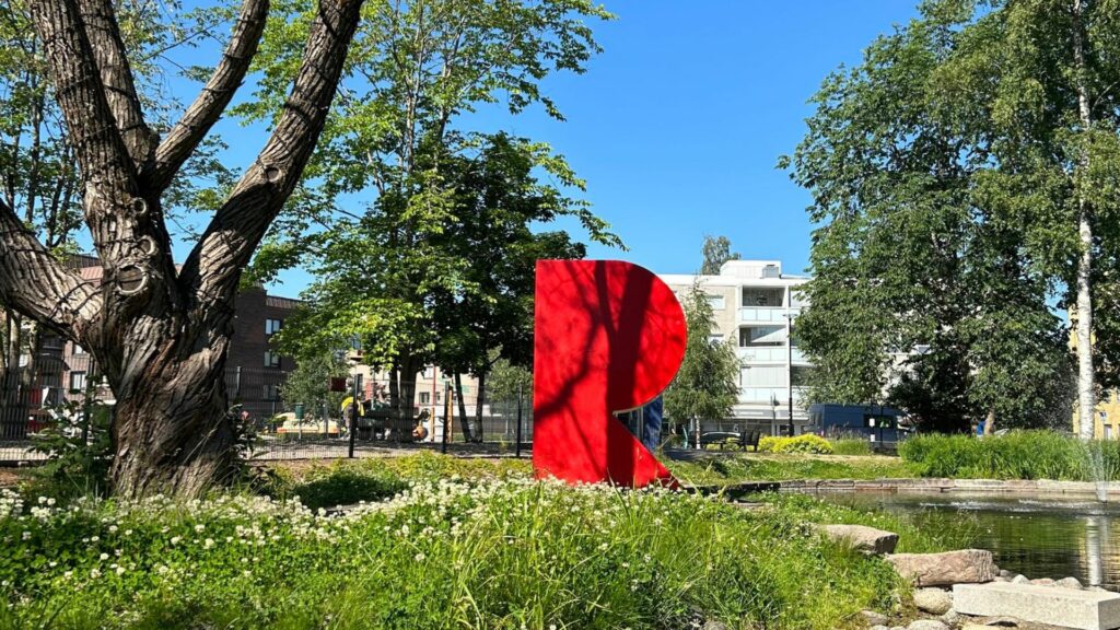 Punainen R-kirjaimen muotoinen muovinen veistos vehreässä puistossa. Taustalla kerrostalo.