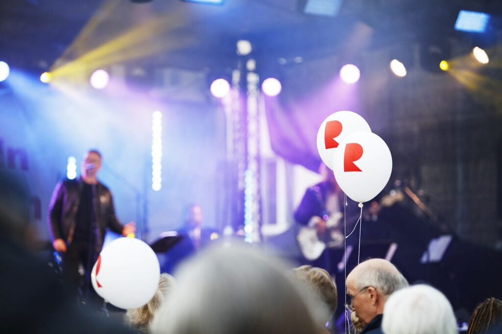 Konserttilava, jossa leijailee sininen sumu, jota halkoo kirkkaat spottivalot. Lavalla esiintyjä. Etualalla yleisön joukosta nousee kaksi ilmapalloa, joissa punainen R-tunnus.
