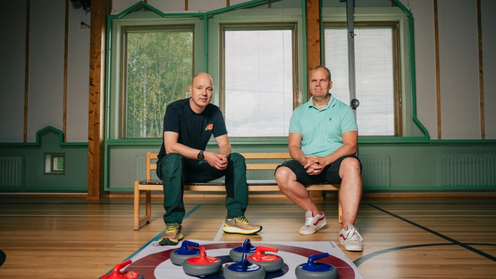 Kaksi urheilullisen näköistä miestä istuu salissa puupenkillä edessään lattialla curling-välineitä.