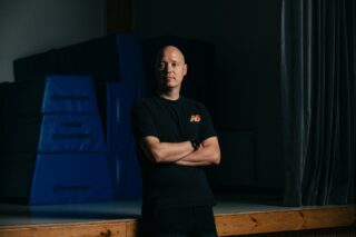 Mustaan t-paitaan pukeutunut mies seisoo kädet puuskassa liikuntasalissa, tumma tausta.
