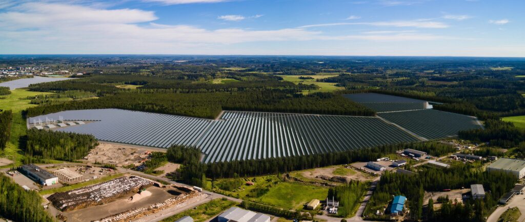 Viistoilmakuvassa aurinkopaneeleja ja ympäröiviä metsä- ja teollisuusalueita.