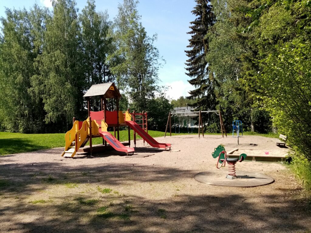 Pellavapuiston nykyiset leikkivälineet: etualalla vihreä lohikäärmejousikiikku, vasemmalla punakeltainen liukumäkikiipeilyteline ja takana keinut.