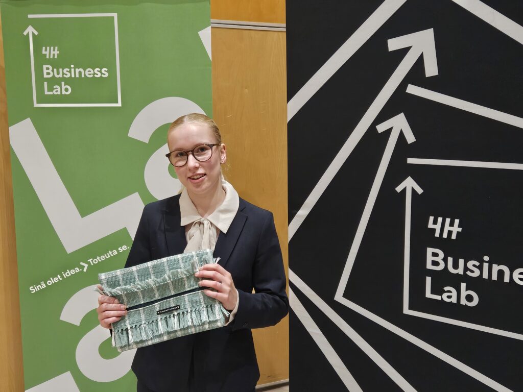 Nuori nainen esittelee kierrätetystä tekstiilistä tekemäänsä vaatekappaletta. Taustalla on 4H:n toimintaa esitteleviä roll uppeja.